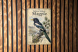 A vintage-style metal sign with a black and white illustration of a magpie perched on a branch, mounted on a wooden wall.
