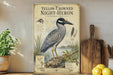 A vintage-style illustration of a yellow-crowned night-heron, a coastal bird species, is displayed on a shelf next to a wooden cutting board and a potted plant.