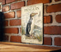 A vintage-style illustration of a yellow-eyed penguin is displayed on a brick wall, with a wooden surface below it.