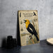 a vintage-style metal plate with an illustration of a yellow-headed blackbird, surrounded by various natural elements such as grass, plants, and a bird feeder.