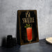 a framed cocktail illustration with a swirled drink and a green leaf garnish, placed on a gray countertop next to a stack of black containers.