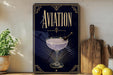 a decorative sign with the word "AVIATION" prominently displayed, along with an illustration of a martini glass and a cherry. The sign is placed on a wooden surface, with a potted plant, a cutting board, and some lemons nearby.