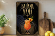 A decorative sign with the text "BAHAMA MAMA" and an illustration of a colorful drink, placed on a wooden surface next to a potted plant and a cutting board.