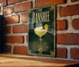 A framed poster of a cocktail with a lemon twist, hanging on a brick wall.