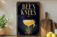 A decorative sign for "Bee's Knees" cocktail, featuring a glass of yellow liquid and a lemon twist, is displayed on a wooden shelf next to a wooden cutting board and a potted plant.