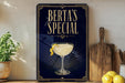 A framed poster with the text "BERTA'S SPECIAL" and an illustration of a cocktail glass with a lemon twist, placed on a wooden shelf next to a cutting board and a potted plant.