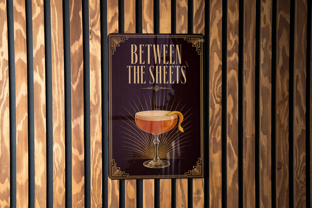 A sign with the text "BETWEEN THE SHEETS" and an illustration of a cocktail glass with a lemon peel.