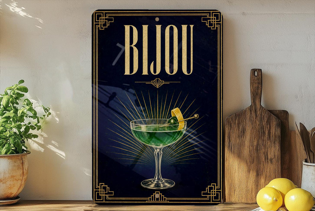 A decorative sign with the word "Bijou" displayed prominently, featuring an illustration of a cocktail glass with a lemon twist. The sign is placed on a wooden surface, with a potted plant, a wooden cutting board, and some lemons nearby.