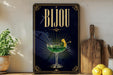 A decorative sign with the word "Bijou" displayed prominently, featuring an illustration of a cocktail glass with a lemon twist. The sign is placed on a wooden surface, with a potted plant, a wooden cutting board, and some lemons nearby.