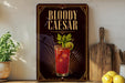 A framed poster of a BloodyCAesar cocktail is displayed on a wall, with a wooden cutting board and a potted plant to the side.