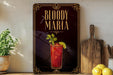 A framed poster advertising "Bloody Maria" cocktail, featuring a red drink with a garnish and a lemon slice, along with a wooden cutting board and a potted plant.