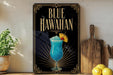 A framed advertisement for a Blue Hawaiian cocktail, featuring a blue drink with a straw and a yellow umbrella, placed on a wooden surface next to a wooden cutting board and a potted plant.
