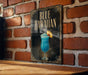 A sign with the text "BLUEHAWAIAN" and an illustration of a blue cocktail with a straw and a fan on a brick wall.