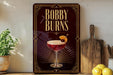A decorative sign for "Bobby Burns" cocktail, featuring an illustration of a cocktail glass with a cherry on top, placed on a wooden surface next to a cutting board and some lemons.