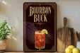 A decorative sign for "Bourbon Buck", featuring an illustration of a cocktail with a lemon slice, placed on a wooden surface next to a potted plant and a cutting board.