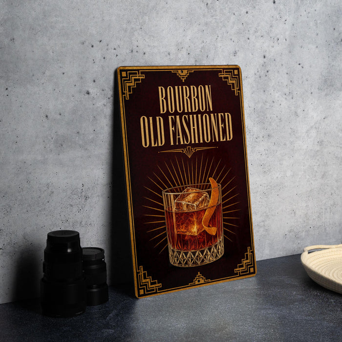 A wooden sign with the text "BOURBON OLD FASHIONED" and an illustration of a cocktail glass with an orange peel on it, placed on a gray countertop next to some black camera lenses.