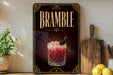 a decorative sign for a drink called "Bramble" with a glass of the drink and garnish, placed on a wooden surface next to a cutting board and some lemons.