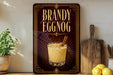 A framed advertisement for "Brandy Eggnog" is displayed on a wall, featuring an illustration of a glass of eggnog with a cinnamon stick.