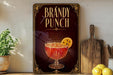 A framed advertisement for "Brandy Punch" featuring a glass of red-colored liquid with a slice of orange on the rim, placed on a wooden surface next to a wooden cutting board and a potted plant.