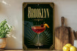 A decorative sign for "Brooklyn" featuring a cocktail glass with a cherry and lemon slice, placed on a wooden surface next to a cutting board and a potted plant.