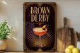 A framed poster of a cocktail called "Brown Derby" is displayed on a wall, with a wooden cutting board and a potted plant to the side.