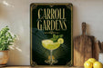 A framed poster advertising Carroll Gardens, featuring a cocktail glass with a lemon slice and mint leaves, is displayed on a shelf next to a wooden cutting board and a potted plant.
