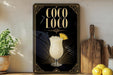 A decorative sign for a coconut drink called "Coco Loco" is displayed on a wooden shelf, accompanied by a potted plant, a cutting board, and some lemons.