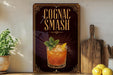 a sign with the text "Cognac Smash" and an illustration of a cocktail, placed on a wooden surface next to a cutting board and some lemons.