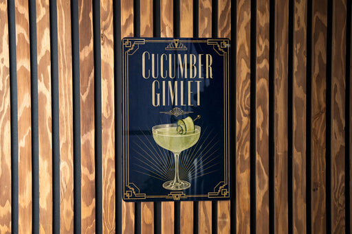 A sign for a cucumber gimlet cocktail is displayed on a wooden wall.
