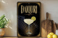 a decorative sign for a Daiquiri cocktail, with a glass of the cocktail and a lemon slice displayed on it. The sign is placed on a wooden surface, with a potted plant and a cutting board nearby.