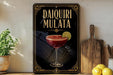 a decorative sign with the text "DAIQUIRI MULATA" and an illustration of a cocktail glass with a lime slice. The sign is placed on a wooden surface, with a potted plant, a wooden cutting board, and some lemons nearby.