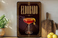 A framed poster of a cocktail called Eldorado, featuring a glass of the drink with a lemon twist, is displayed on a wall next to a wooden cutting board and some kitchen utensils.