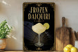 A framed advertisement for a frozen daiquiri cocktail, featuring a glass of the drink and a lime slice, is displayed on a wall next to a wooden cutting board and a potted plant.