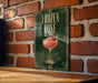 A framed poster with the title "FROZEN ROSE" and an illustration of a pink drink in a glass, hanging on a brick wall.