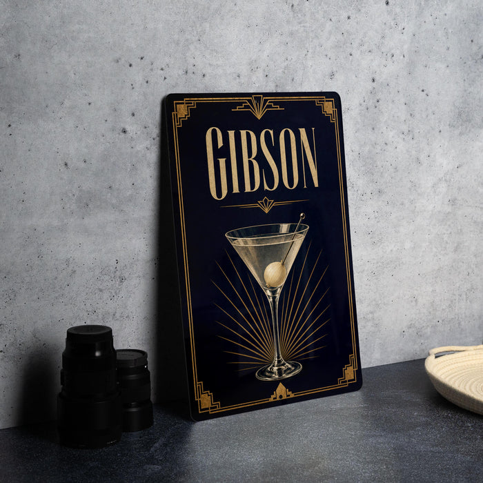 A framed poster of a martini glass with the word "GIBSON" on it, placed on a gray countertop next to some black camera lenses.