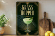 A framed poster with the title "GRASS HOPPER" displayed prominently, featuring an illustration of a green cocktail with a garnish. The poster is set against a dark background with a decorative border, and there are some potted plants and kitchen utensils visible in the foreground.