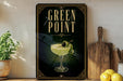 A framed poster with the text "GREEN POINT" and an illustration of a cocktail glass with a lemon slice on it, placed on a wooden shelf next to a wooden cutting board and a potted plant.