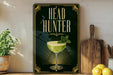 a decorative sign with the text "HEAD HUNTER" and an illustration of a cocktail glass with a mint leaf garnish. The sign is displayed on a shelf next to a wooden cutting board and a potted plant.