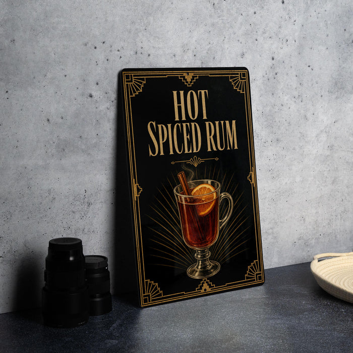 a black sign with a decorative border, displaying an illustration of a hot spiced rum drink with a slice of lemon and a cinnamon stick.
