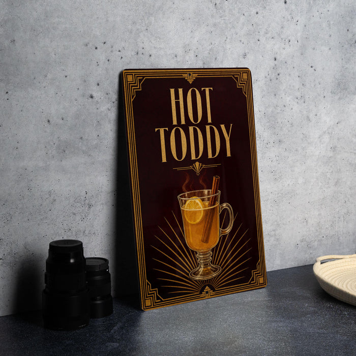 A framed advertisement for "Hot Toddy" featuring a glass mug with a slice of lemon and a cinnamon stick, placed on a gray countertop next to a stack of black containers.