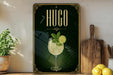 A decorative sign for a drink called "HUGO" featuring a glass of lemonade with a mint garnish, placed on a wooden surface next to a wooden cutting board and a potted plant.