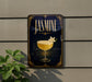 A decorative sign with the word "JASMINE" prominently displayed, featuring an illustration of a glass filled with a yellow liquid and a flower.