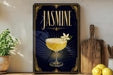 A framed poster with a glass of jasmine cocktail and a white flower on a dark background, placed on a wooden shelf next to a wooden cutting board and a potted plant.