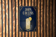 A poster for a cocktail called "John Collins" is displayed on a wooden wall.