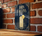 A sign for John Collins cocktail is displayed on a brick wall, featuring an illustration of a cocktail glass with a lemon slice.