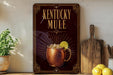 A decorative sign for Kentucky Mule, featuring a copper mug with a lemon slice, is displayed on a shelf next to a wooden cutting board and a potted plant.