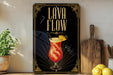 A framed poster with the text "Lava Flow" and an illustration of a colorful drink, placed on a wooden shelf next to a wooden cutting board and a potted plant.