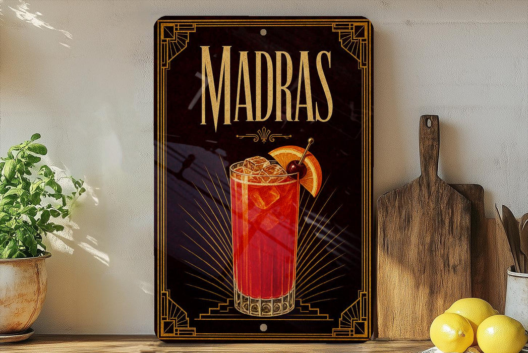 A decorative sign for Madras cocktails, featuring an illustration of a red cocktail with a lemon slice and a cherry on top, is displayed on a shelf next to a wooden cutting board and a potted plant.