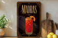 A decorative sign for Madras cocktails, featuring an illustration of a red cocktail with a lemon slice and a cherry on top, is displayed on a shelf next to a wooden cutting board and a potted plant.