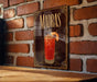 A framed poster of a Madras cocktail hangs on a brick wall, featuring an illustration of the drink and decorative elements.